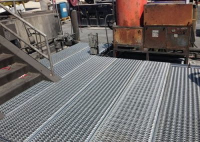 Metal grate flooring near industrial machinery and a staircase, with various residential HVAC equipment and structures in the background.