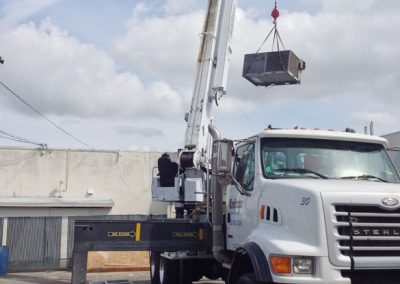 A crane truck hoisting a residential HVAC unit in an urban environment with wires and a building in the background, set against the vibrant backdrop of Orange County, California.